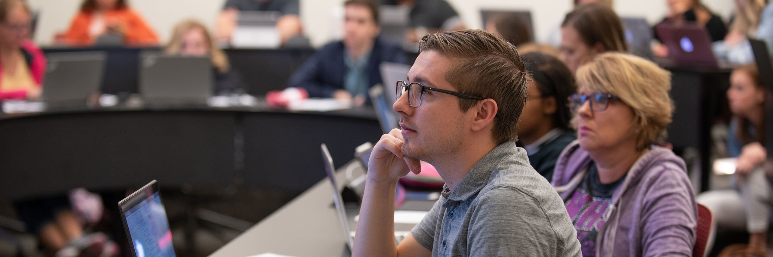 Professors and Staff in a classroom, attentively listening with laptops open.